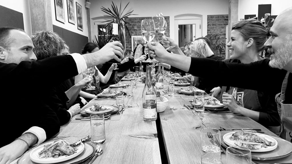 Zu sehen ist das gesamte CUBE-Team an einer großen Tafel beim Anstoßen mit Wein.