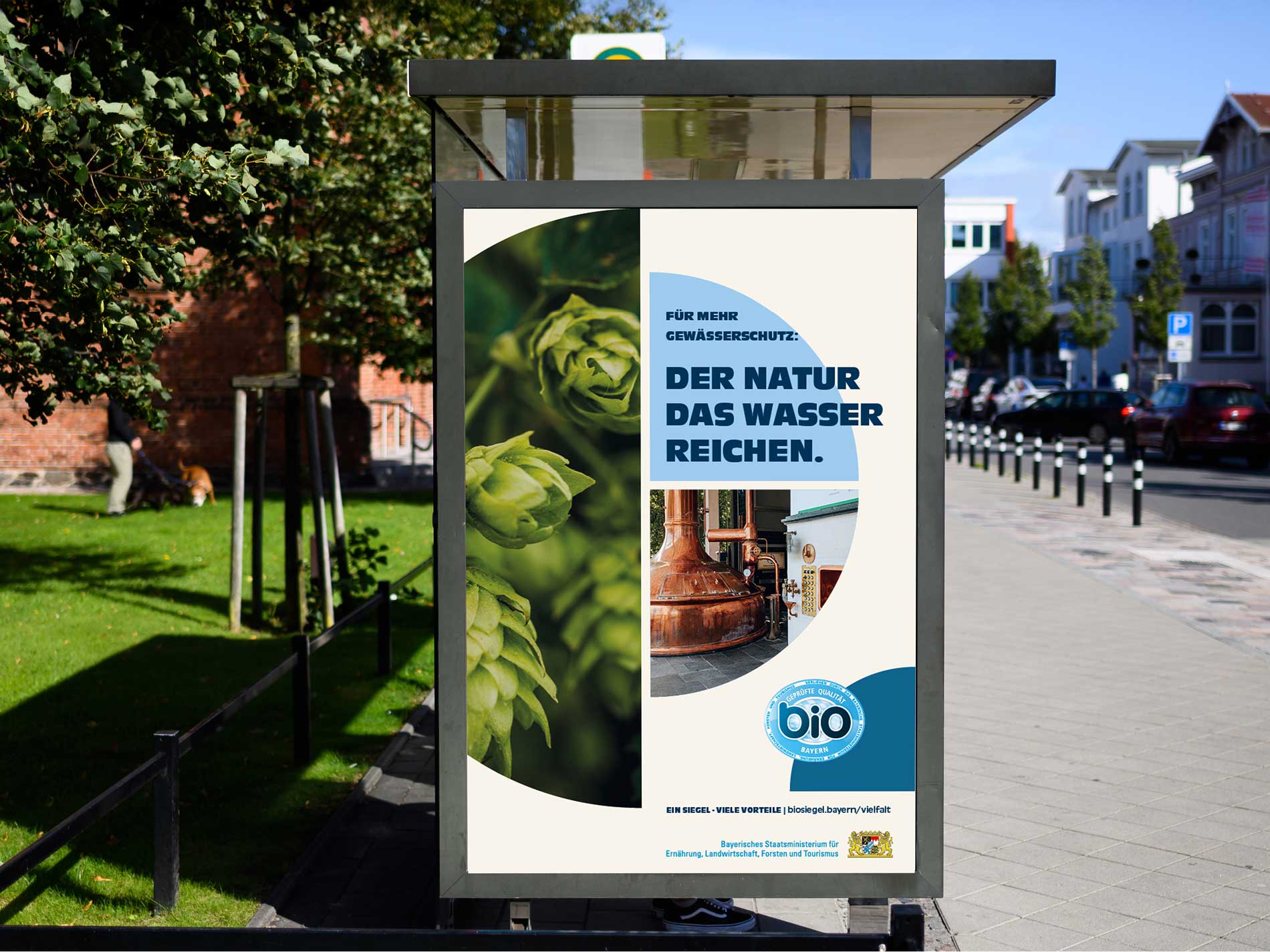 Plakat an einer Bushaltestelle mit einer Kampagne für das Bayerische Bio-Siegel. Es zeigt Hopfen, eine Brauerei und den Slogan „Für mehr Gewässerschutz: Der Natur das Wasser reichen.“