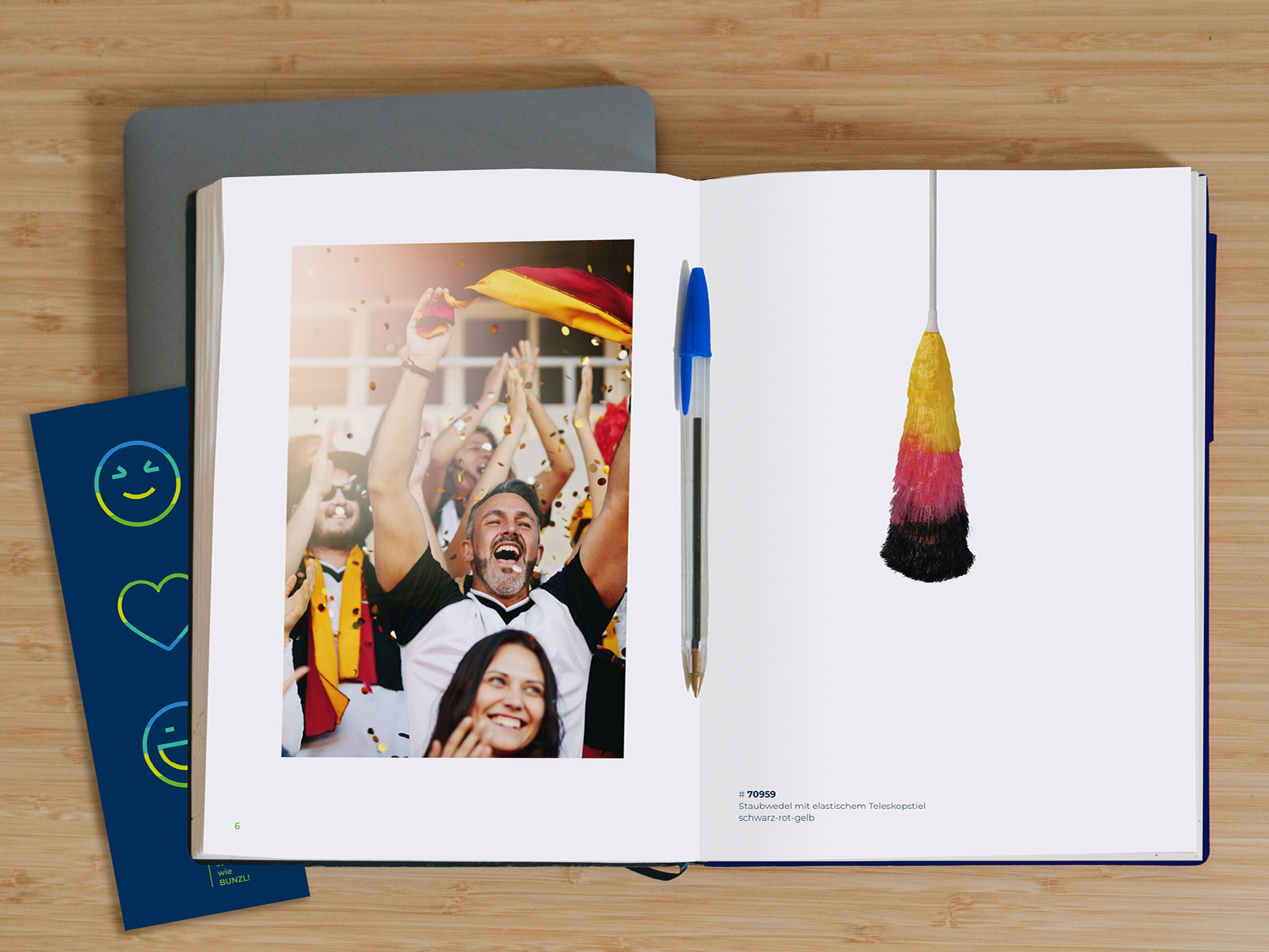Aufgeschlagenes Notizbuch auf einem Holztisch, daneben ein blauer Kugelschreiber. Links im Buch ein Foto jubelnder Fußballfans in Deutschlandfarben, rechts ein Staubwedel in Schwarz-Rot-Gelb mit der Artikelnummer und Beschreibung darunter.