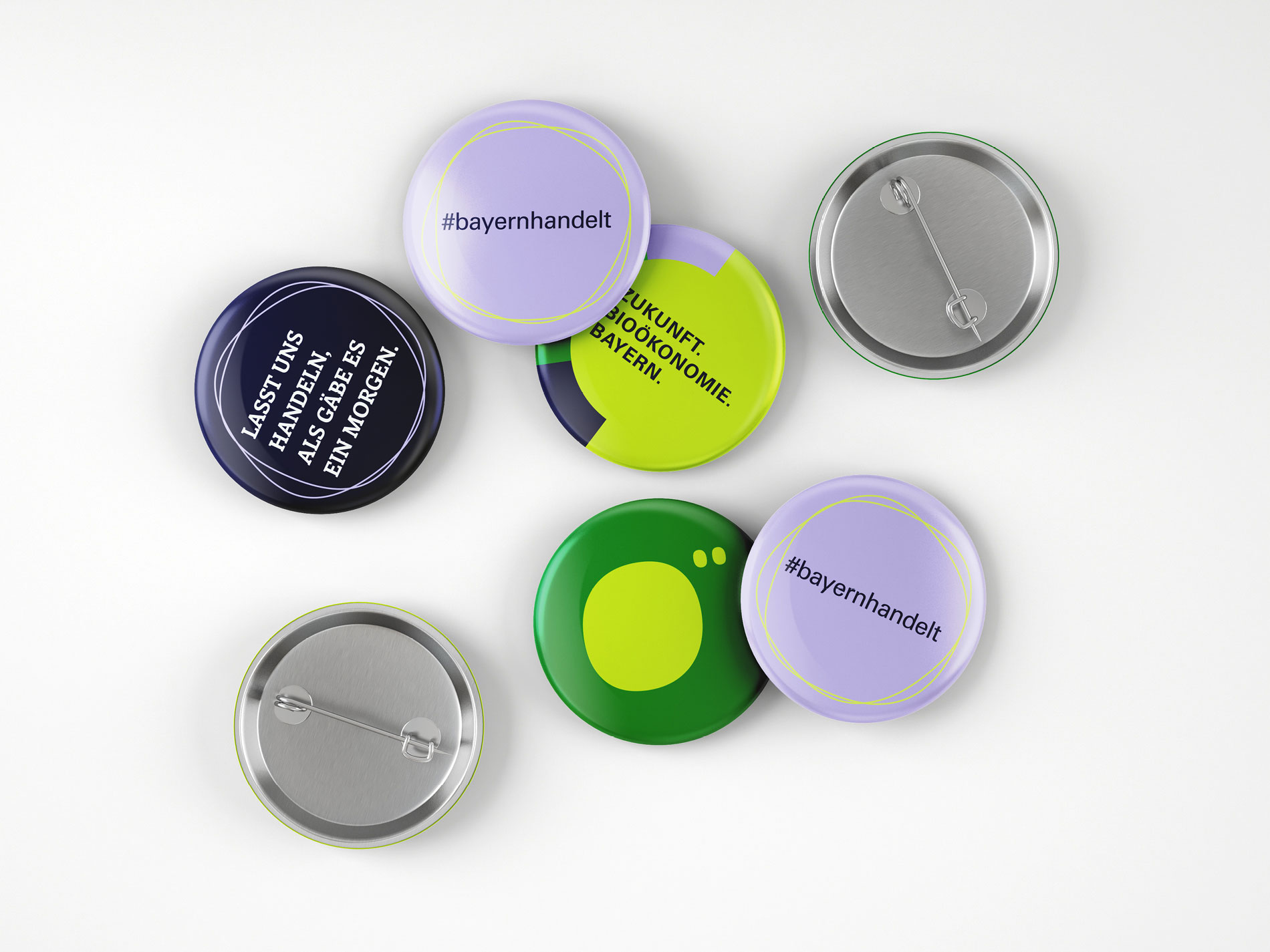 Das Bild zeigt mehrere bunte Buttons mit verschiedenen Aufschriften wie "#bayernhandelt" und "Lasst uns handeln, als gäbe es ein Morgen", die auf einer weißen Oberfläche liegen.