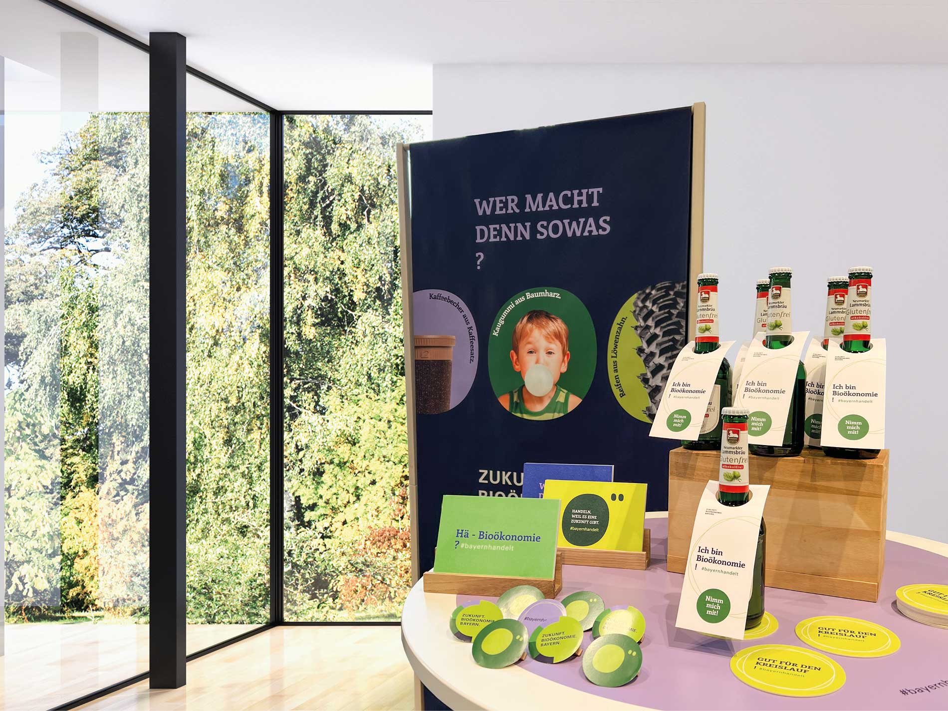 Das Bild zeigt einen Ausstellungstisch mit Bioökonomie-Produkten wie Bierflaschen mit dem Etikett "Ich bin Bioökonomie" und Informationsmaterialien, begleitet von einem Poster im Hintergrund, das nachhaltige Produkte erklärt.