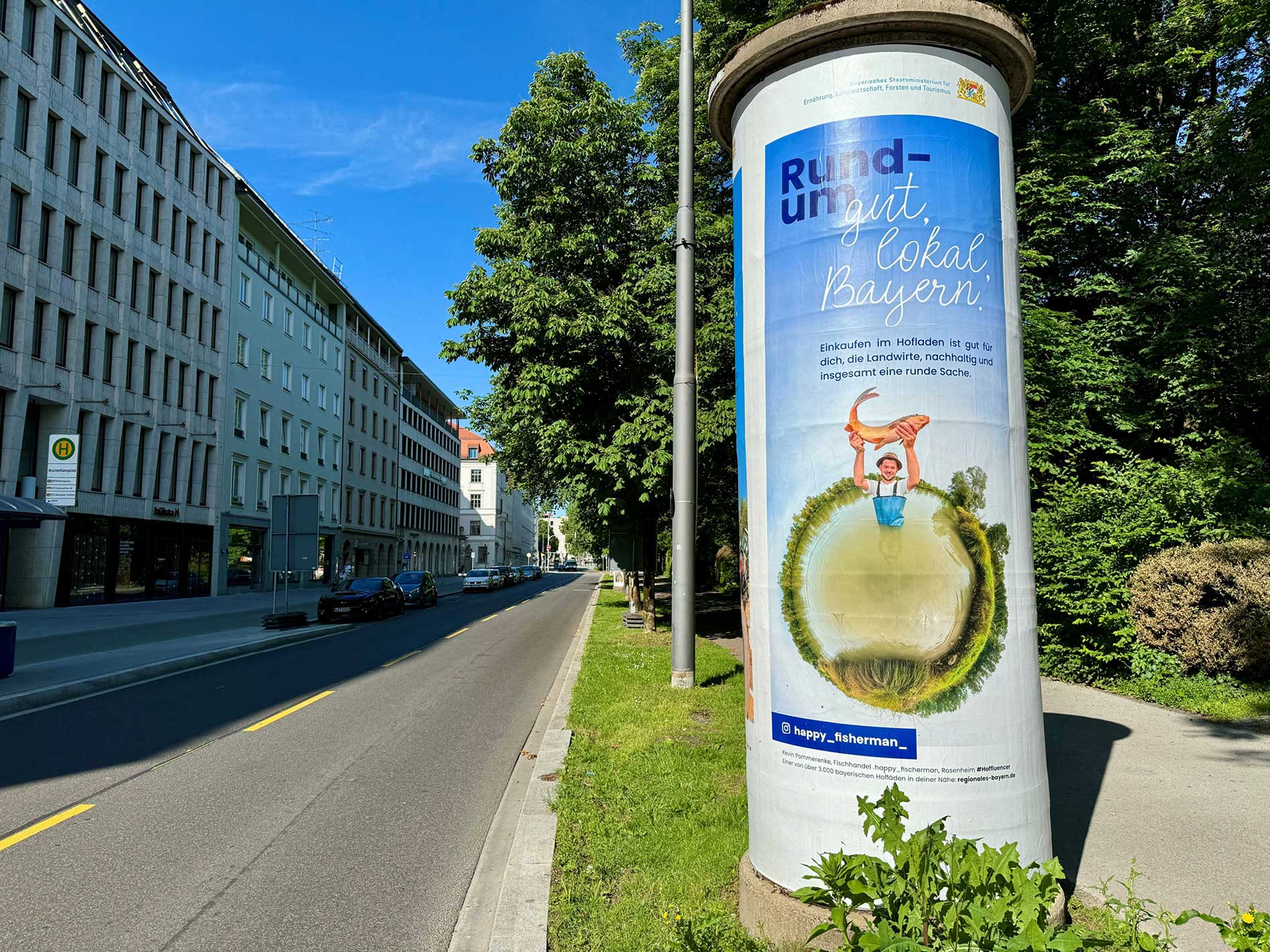 Das Bild zeigt eine Litfaßsäule am Straßenrand, die mit einem Plakat der Direktvermarkter Kampagne "Rund um gut, lokal, Bayern" beklebt ist, das für lokale Produkte und den Einkauf im Hofladen wirbt.