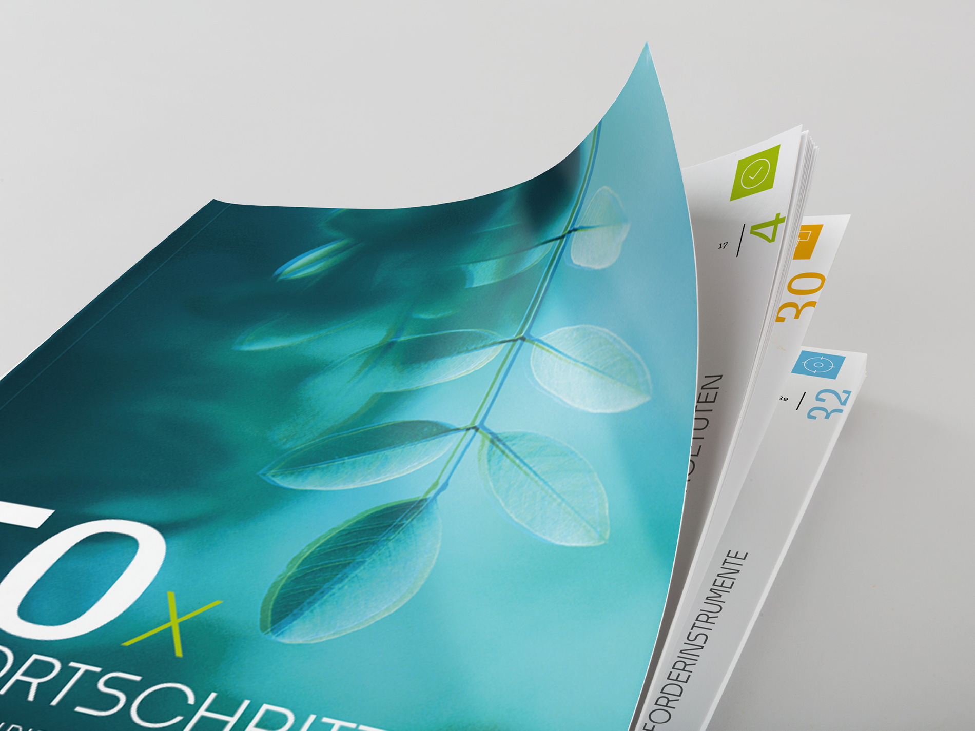 Das Bild zeigt den teilweise umgeblätterte Fortschrittsbericht mit einem grünen, pflanzenartigen Cover und nummerierten Seiten, die verschiedene Abschnitte darstellen.