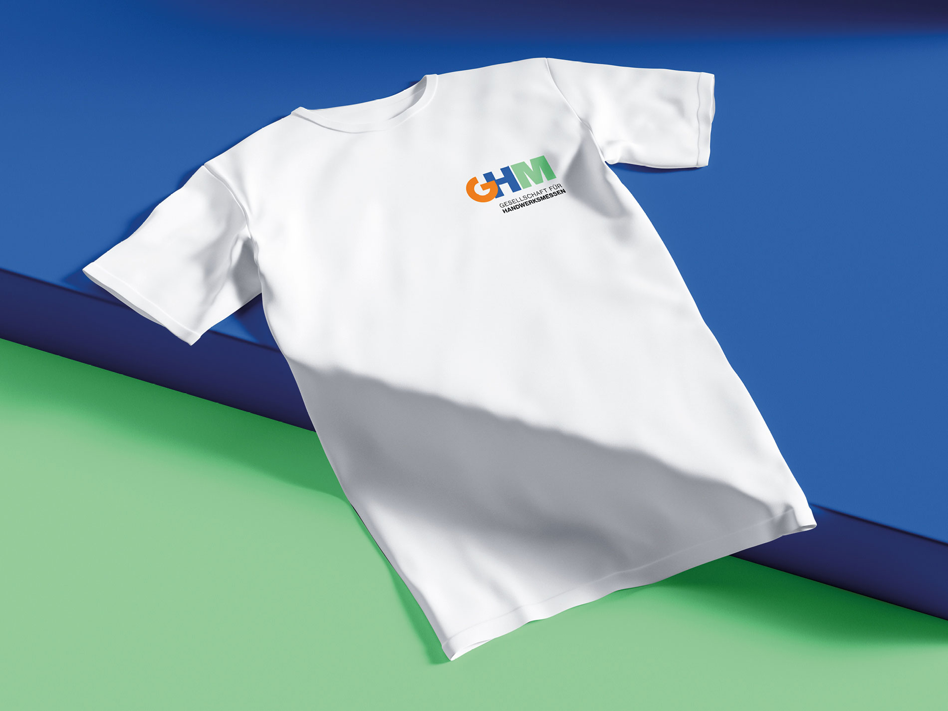 Das Bild zeigt ein weißes T-Shirt, auf dem das Logo der "Gesellschaft für Handwerksmessen" (GHM) auf der Brust aufgedruckt ist. Das T-Shirt liegt auf einer Oberfläche, die in Blau und Grün gestaltet ist. Das GHM-Logo ist in den Farben Orange, Blau und Grün, ähnlich wie auf den zuvor gezeigten Karten, aufgedruckt.