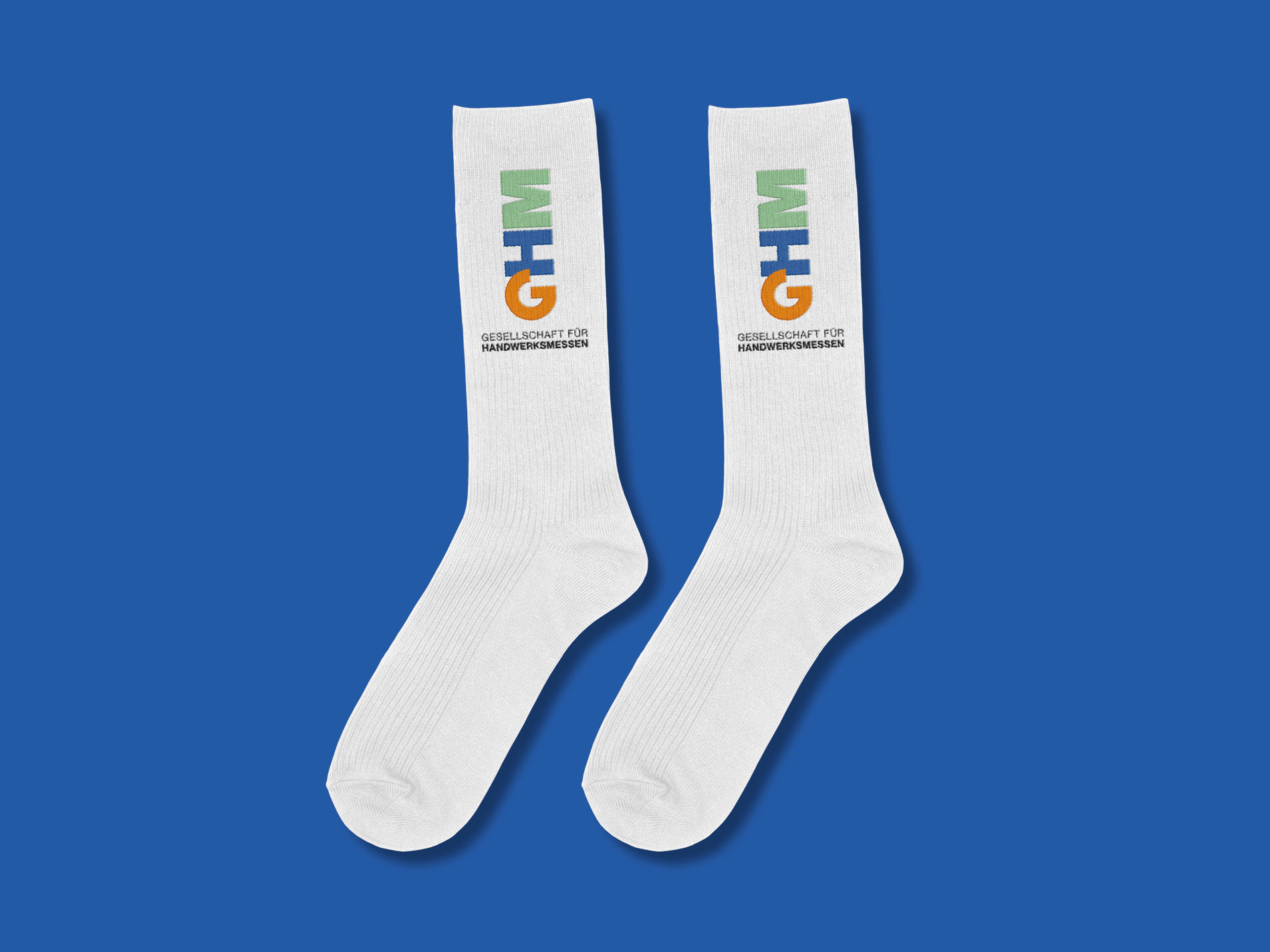 Das Bild zeigt ein Paar weiße Socken, auf denen das Logo der "Gesellschaft für Handwerksmessen" (GHM) in den Farben Orange, Blau und Grün aufgedruckt ist. Die Socken liegen auf einem blauen Hintergrund, und unter dem Logo steht der Schriftzug "Gesellschaft für Handwerksmessen" in Schwarz.