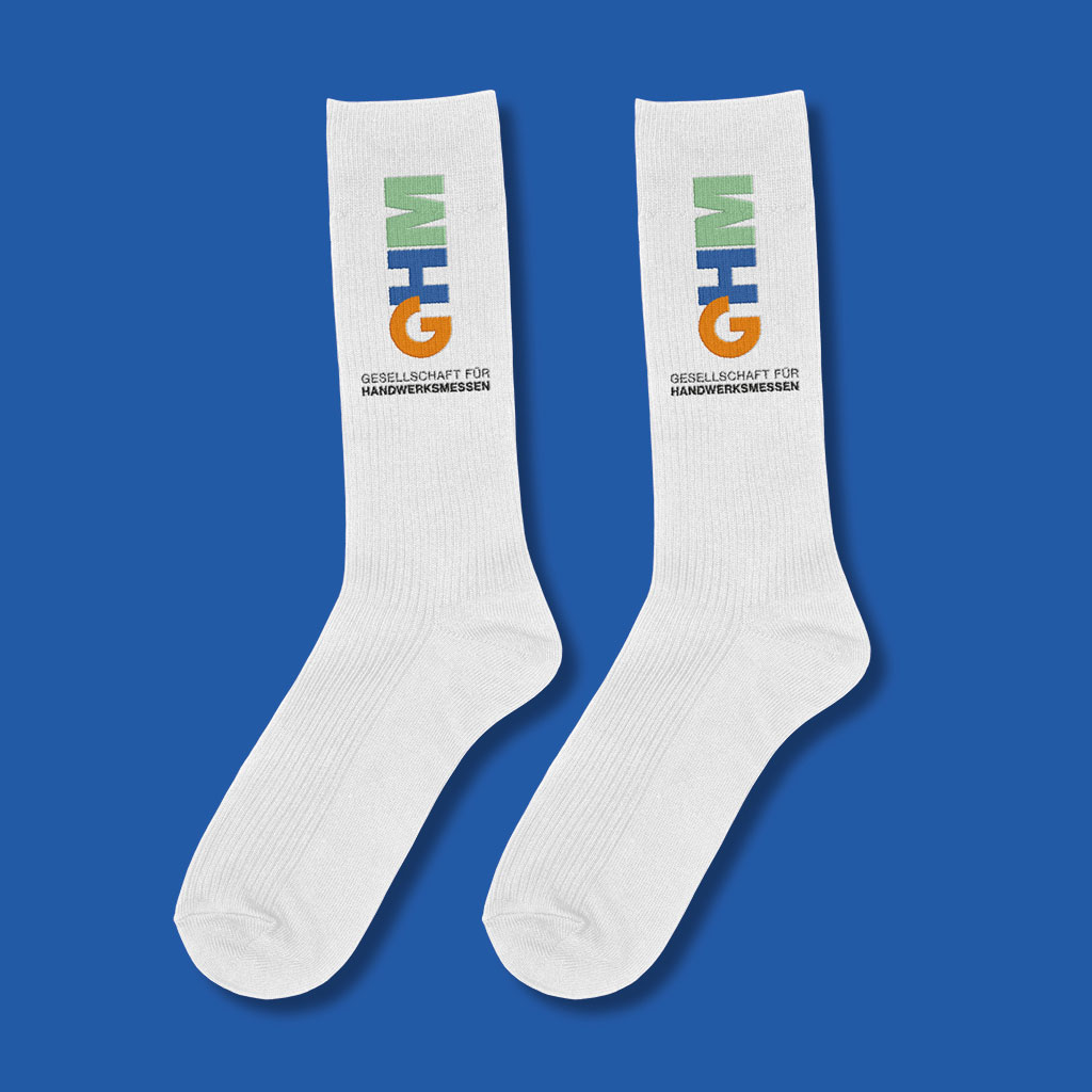 Das Bild zeigt ein Paar weiße Socken, auf denen das Logo der "Gesellschaft für Handwerksmessen" (GHM) in den Farben Orange, Blau und Grün aufgedruckt ist. Die Socken liegen auf einem blauen Hintergrund, und unter dem Logo steht der Schriftzug "Gesellschaft für Handwerksmessen" in Schwarz.