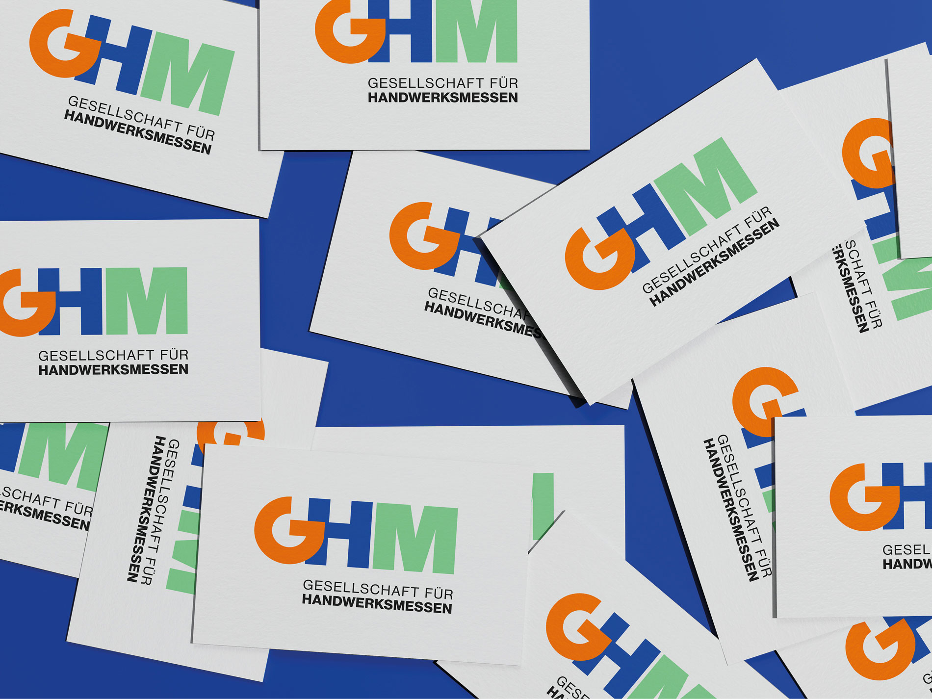 Das Bild zeigt mehrere übereinanderliegende Karten mit dem Logo der "Gesellschaft für Handwerksmessen" (GHM). Das Logo besteht aus den Buchstaben "GHM", die in den Farben Orange, Blau und Grün gestaltet sind, darunter der Schriftzug "Gesellschaft für Handwerksmessen" in Schwarz. Die Karten liegen auf einem blauen Hintergrund verstreut.