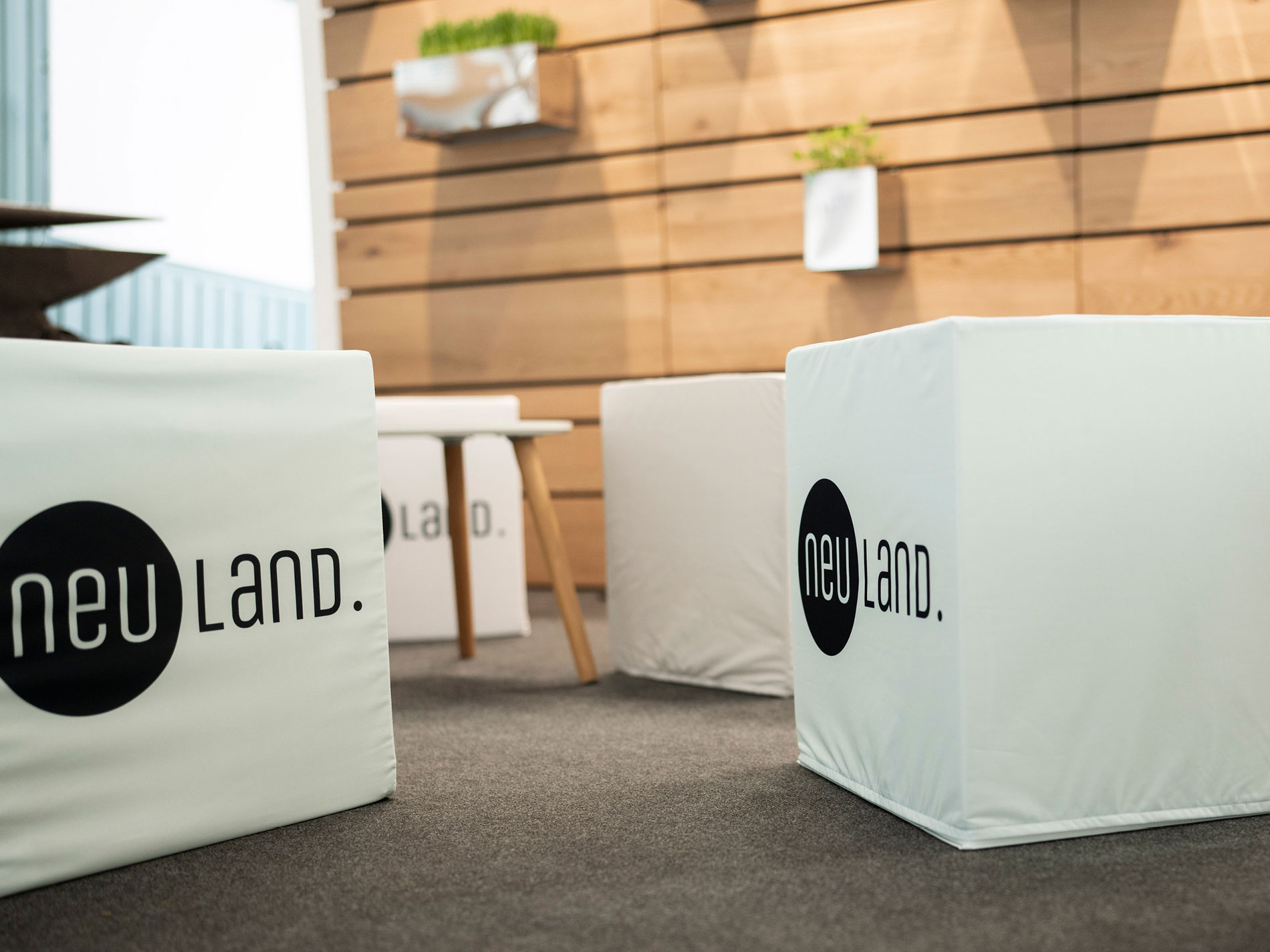 Das Bild zeigt einen modernen Raum mit weißen Sitzwürfeln, auf denen das Logo "neu land." zu sehen ist, sowie einem minimalistischen Holzhintergrund und Pflanzen als Dekoration.