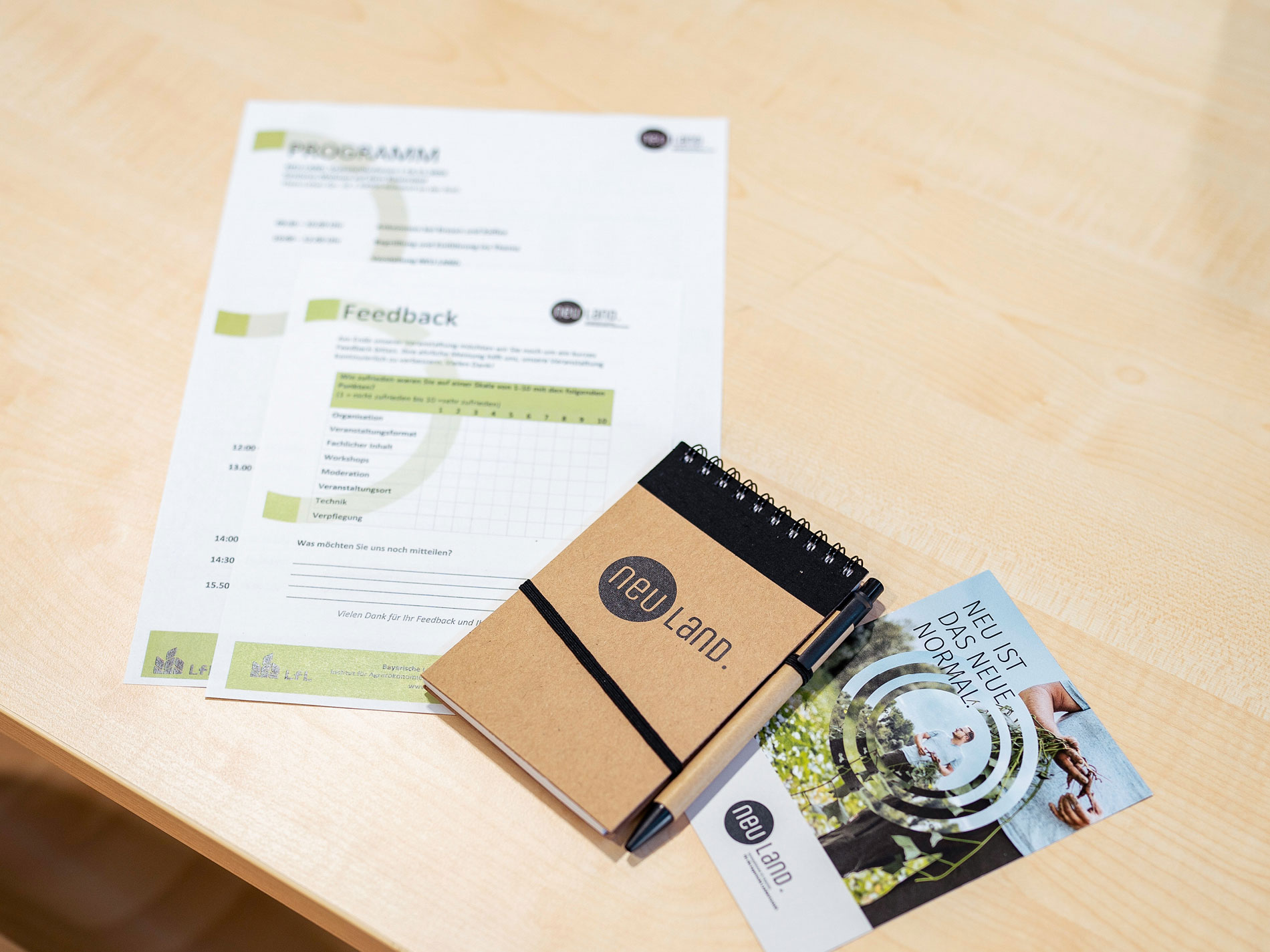 Das Bild zeigt Veranstaltungsunterlagen von Neuland auf einem Tisch, einschließlich eines Programms, eines Feedbackformulars, eines Notizblocks und eines Flyers mit dem Slogan "Neu ist das neue Normal".