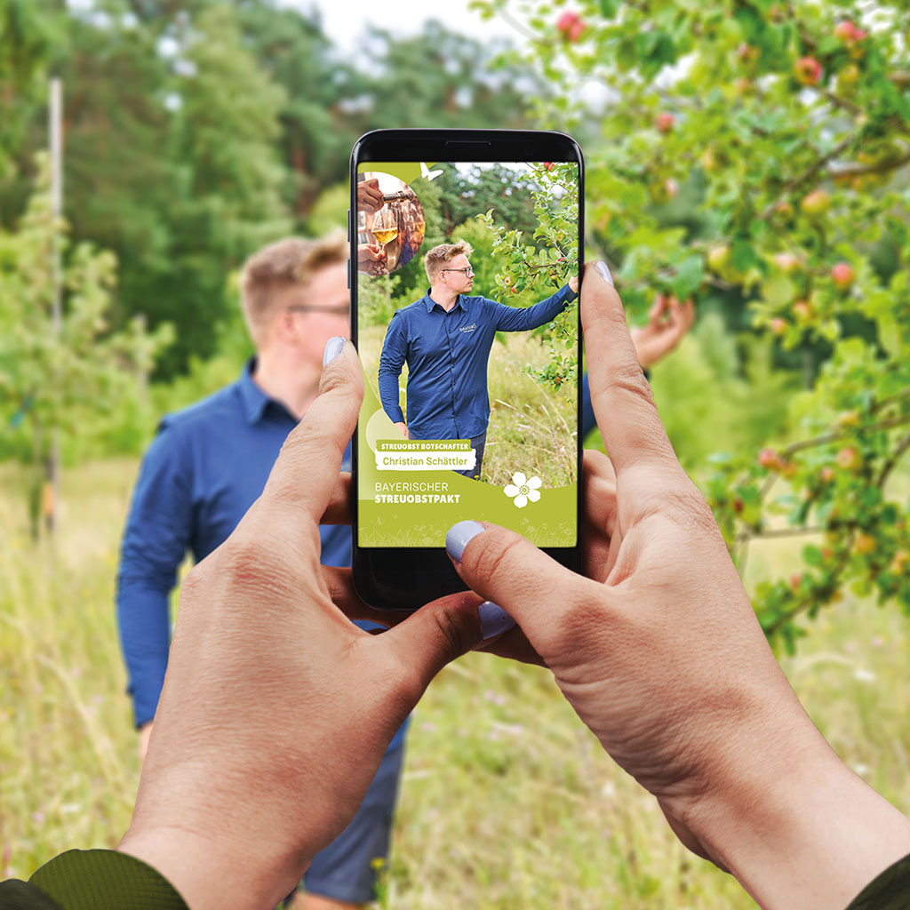 Das Bild zeigt eine Person, die ein Smartphone hält und ein Foto von einem Mann macht, der in einem Obstgarten steht. Der Hintergrund zeigt eine grüne, bewaldete Landschaft mit Apfelbäumen.