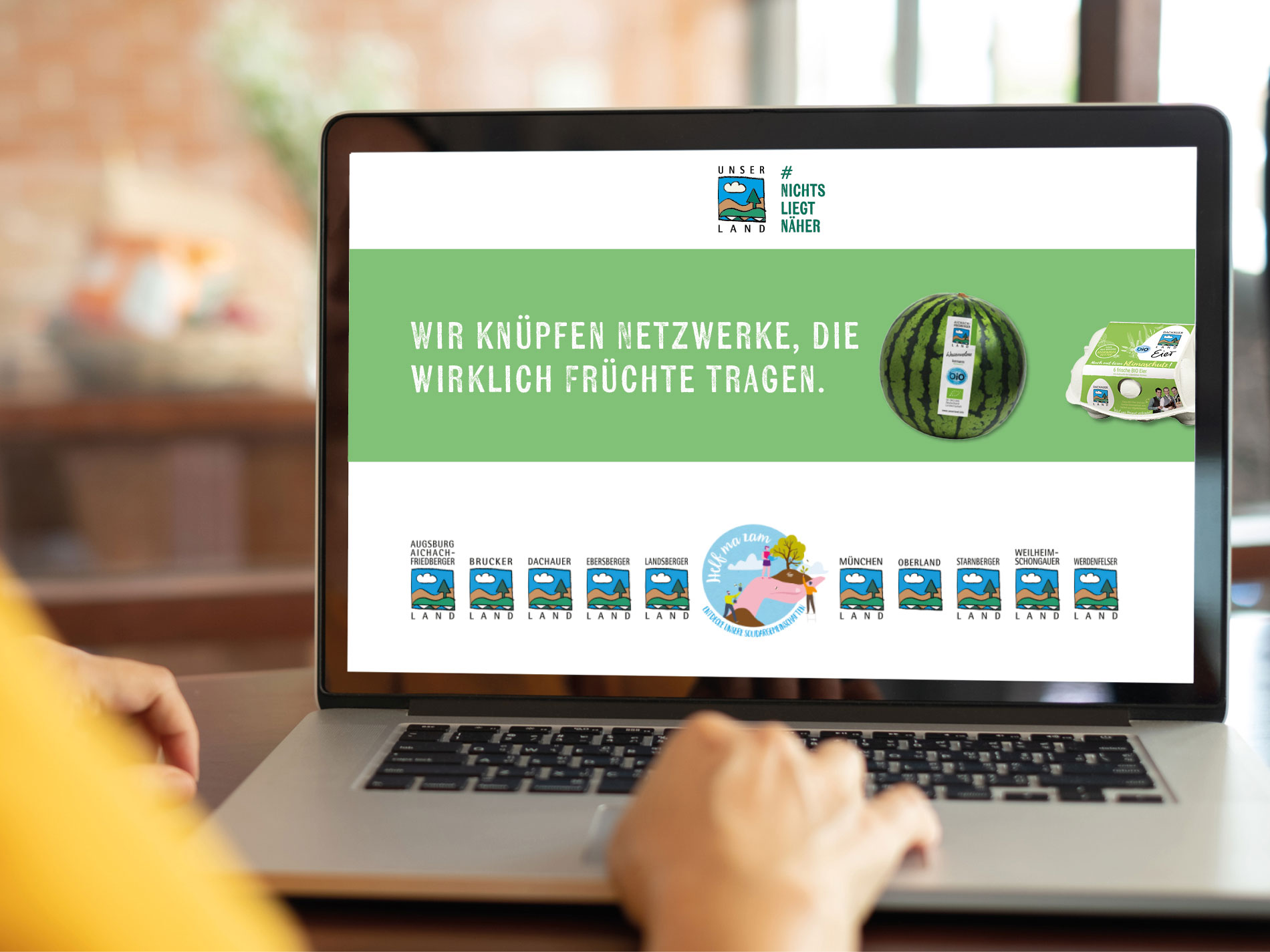 Ein Laptop mit geöffneter Webseite von UNSER LAND. Die Seite zeigt das Logo, verschiedene regionale Partnernetzwerke und den Slogan „Wir knüpfen Netzwerke, die wirklich Früchte tragen“, begleitet von Bildern einer Wassermelone und einem Eierkarton mit Bio-Siegel.