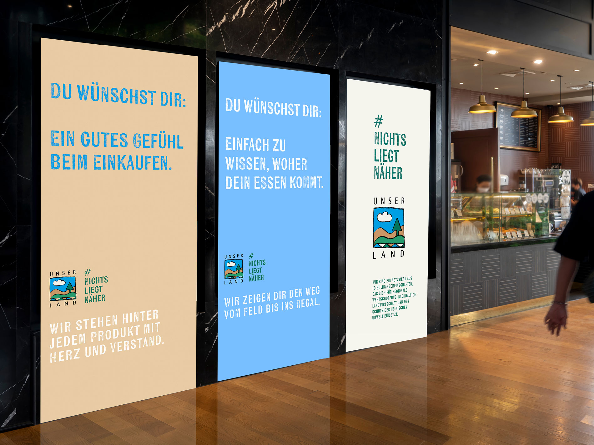 Drei digitale Werbeflächen in einem Einkaufszentrum. Die Anzeigen tragen die Botschaften „Ein gutes Gefühl beim Einkaufen“, „Einfach zu wissen, woher dein Essen kommt“ und „#NICHTS LIEGT NÄHER“, alle mit dem UNSER LAND Logo.