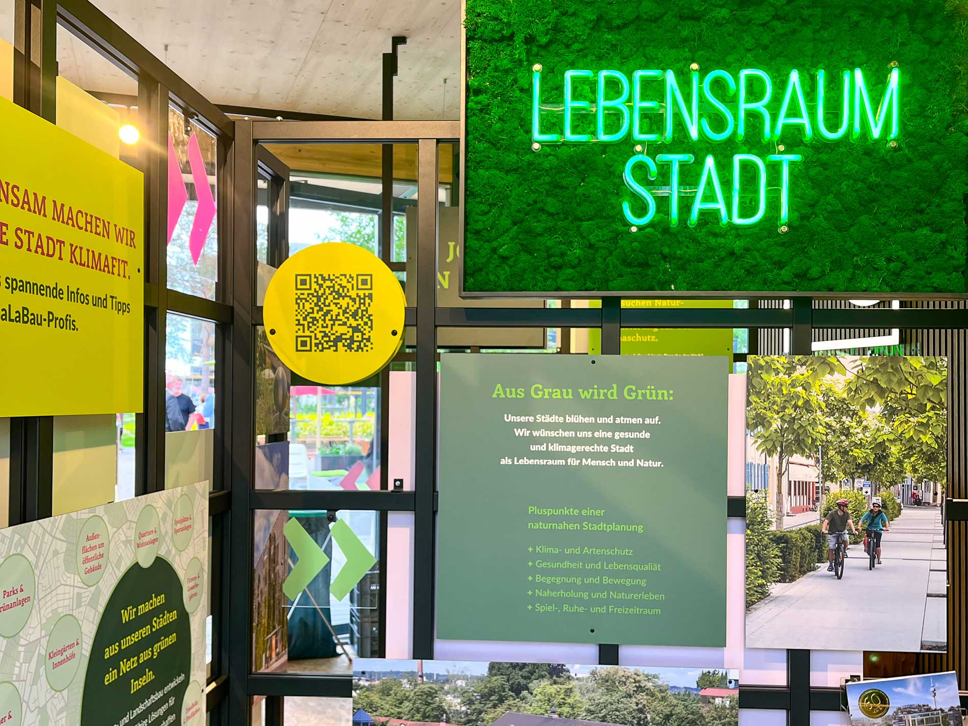 Innenraum des VGL-Pavillons mit dem Thema "Lebensraum Stadt", in der verschiedene Informationstafeln und Schilder mit grünen und gelben Akzenten präsentiert werden, die sich auf klimagerechte Stadtplanung und nachhaltige urbane Entwicklung fokussieren.