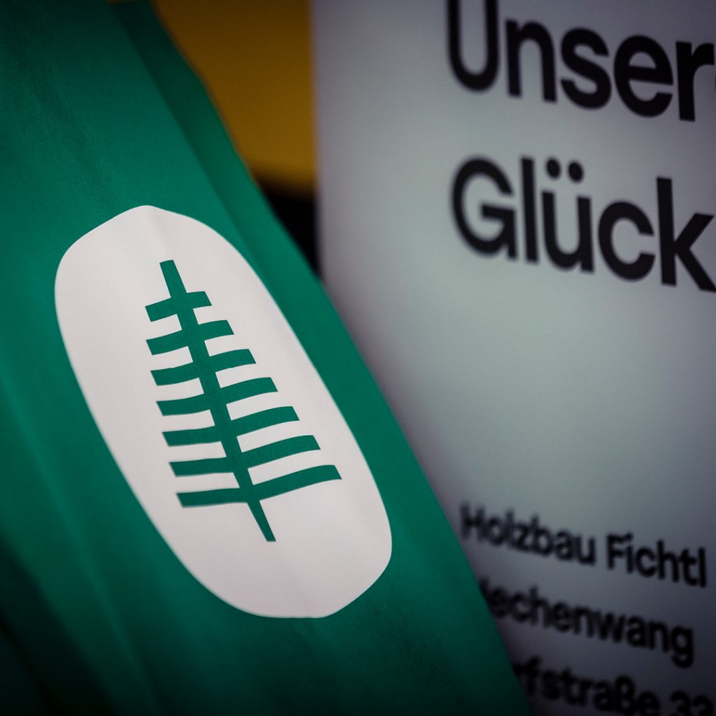 Nahaufnahme einer grünen Stoffflagge mit dem Logo von Holzbau Fichtl – stilisierte weiße Tanne in ovaler Form.