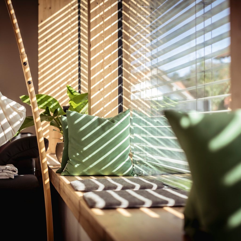 Sonnige Innenraum-Szene mit Holzlamellenwand, Sitzbank aus Holz, grünem Dekokissen und Zimmerpflanze – Lichtspiel durch Jalousien.