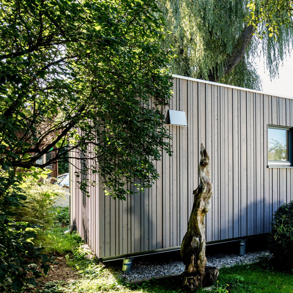 Modernes Holzhaus mit grauer, vertikaler Lattung in einem grünen Garten unter einer Trauerweide, im Vordergrund eine ausgestellte Wurzel als Skulptur.