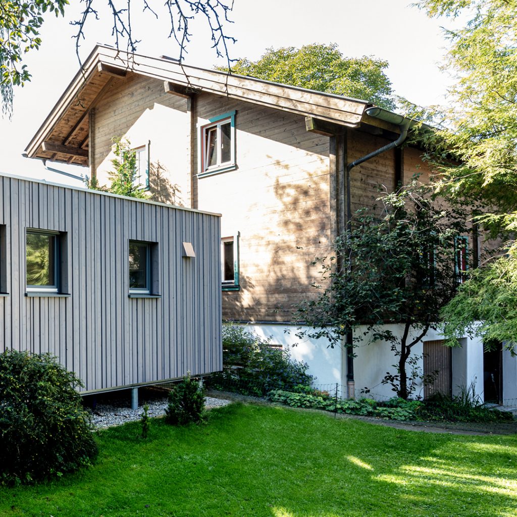 Überdachter Holzbalkon eines Wohnhauses mit Lärchenholzfassade, Holzdielenboden und rustikalem Tisch mit zwei Stühlen, im Hintergrund Blick ins Grüne.