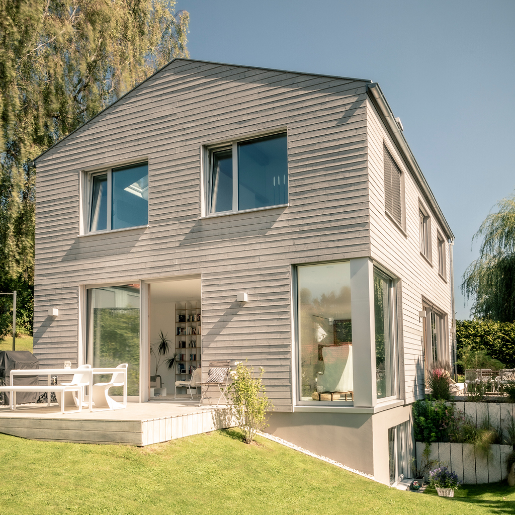 Modernes zweigeschossiges Holzhaus mit silbergrau verwitterter Holzfassade, großen Fensterfronten und Terrasse im Garten.