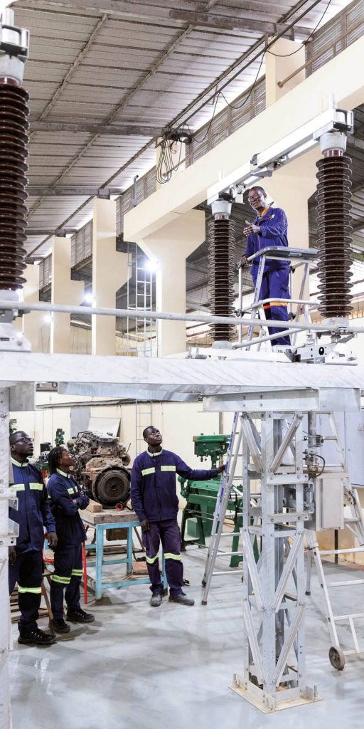 Drei Auszubildende und ein Ausbilder in blauen Arbeitsanzügen in einer Werkhalle mit Hochspannungsanlagen und Maschinen – Reportagefoto aus einem KfW-Entwicklungsprojekt.