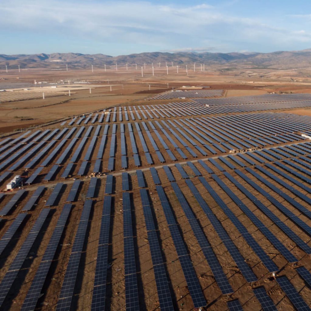 Luftaufnahme einer großen Photovoltaik-Freiflächenanlage in trockener Berglandschaft, im Hintergrund Windräder.