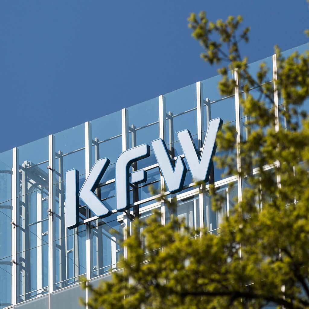 KfW-Logo an einer verglasten Gebäudefassade vor blauem Himmel, im Vordergrund unscharfe Baumzweige.