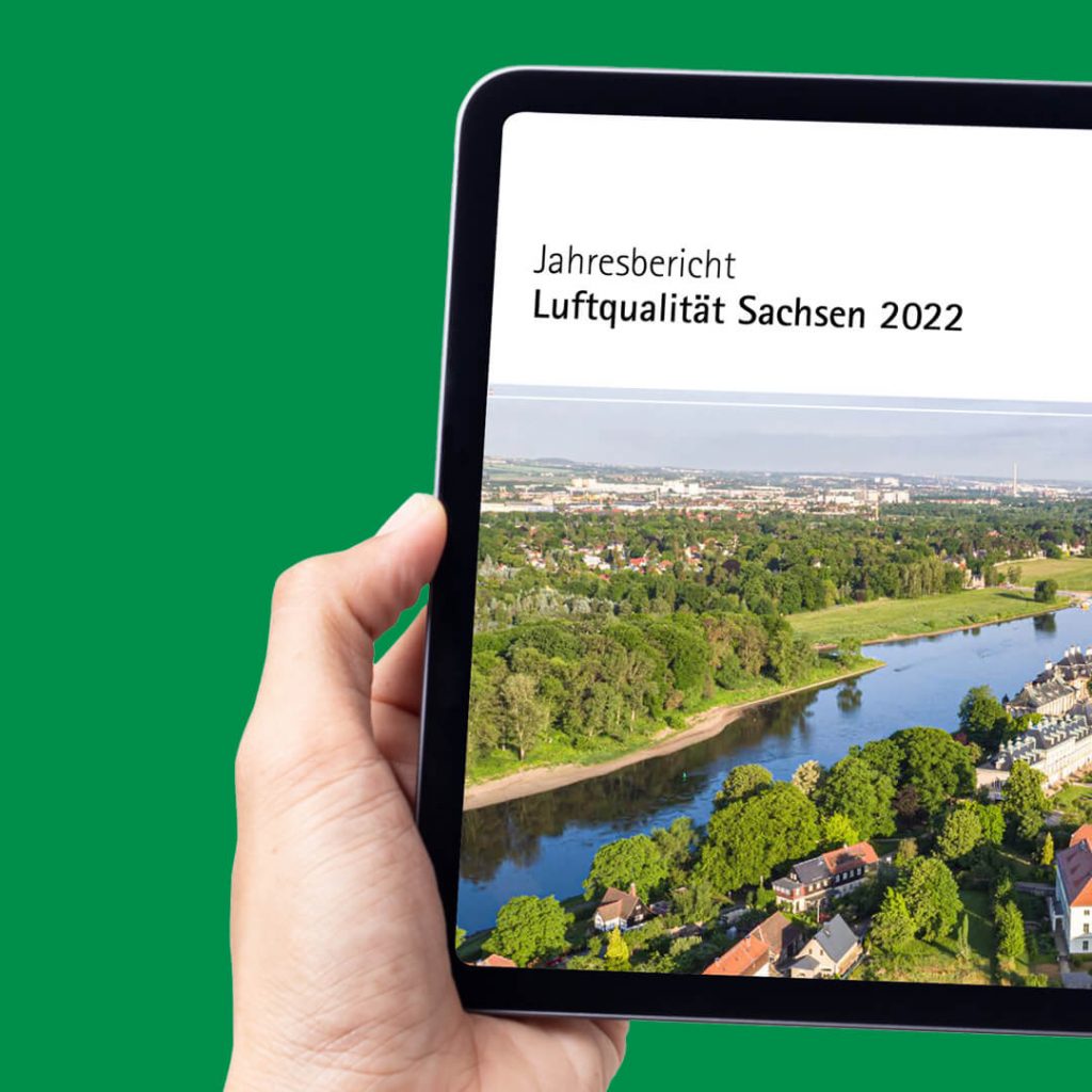 Hand hält ein Tablet mit dem Cover des „Jahresbericht Luftqualität Sachsen 2022" und einer Luftaufnahme der Elbe.